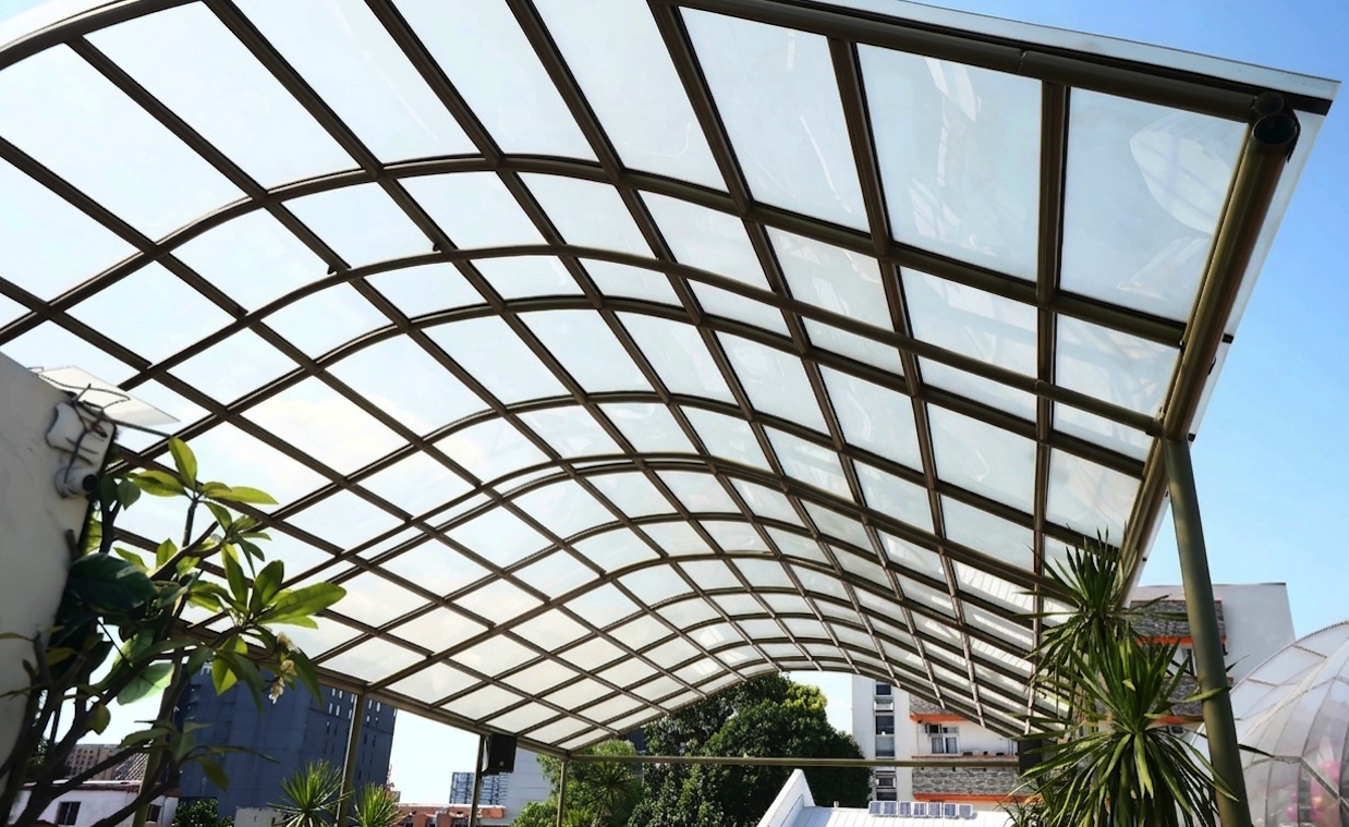 A curved outdoor pergola canopy made of semi-transparent clear polycarbonate sheets installed on a brown metal frame structure against a blue sky.